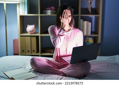 Young Hispanic Woman Using Computer Laptop On The Bed Covering One Eye With Hand, Confident Smile On Face And Surprise Emotion. 