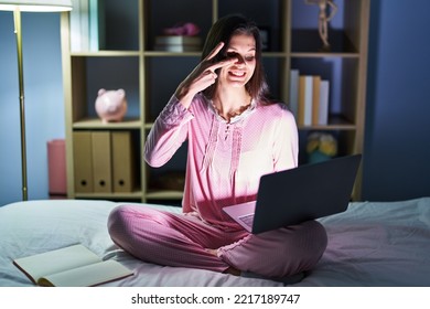 Young Hispanic Woman Using Computer Laptop On The Bed Doing Peace Symbol With Fingers Over Face, Smiling Cheerful Showing Victory 