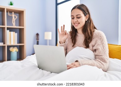 Young Hispanic Woman Using Computer Laptop On The Bed Doing Ok Sign With Fingers, Smiling Friendly Gesturing Excellent Symbol 