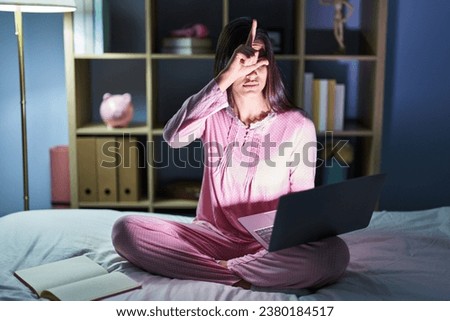 Young hispanic woman using computer laptop on the bed making fun of people with fingers on forehead doing loser gesture mocking and insulting. 