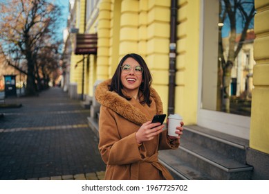 Young Hispanic Woman Using A Smartphone And Drinking Coffee Takeaway While Walking Outside