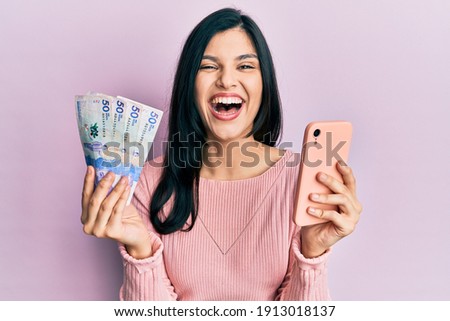 Young hispanic woman using smartphone holding colombian pesos banknotes smiling and laughing hard out loud because funny crazy joke. 