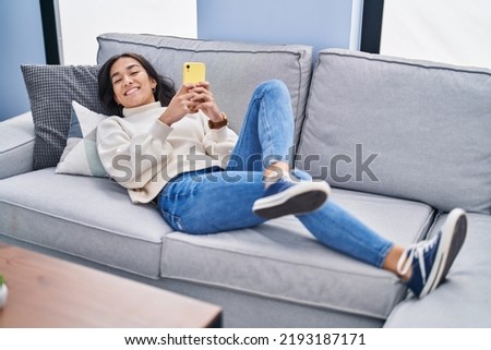 Young hispanic woman using smartphone lying on sofa at home