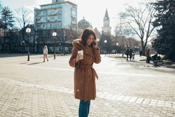 Young Hispanic Woman Walking In The City While Talking On The Smartphone And Drinking Coffee Takeaway