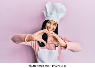 Young hispanic woman wearing baker uniform and cook hat smiling in love showing heart symbol and shape with hands. romantic concept. 