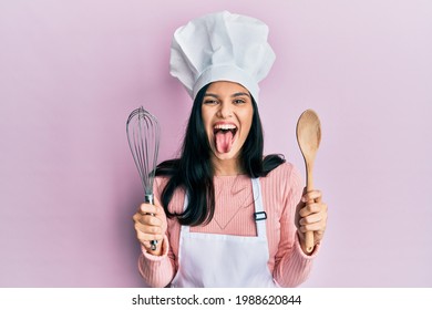 Young hispanic woman wearing baker uniform holding spoon and whisk sticking tongue out happy with funny expression. 