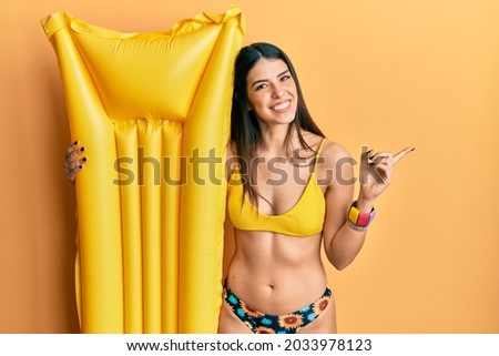 Young hispanic woman wearing bikini and holding summer float smiling happy pointing with hand and finger to the side 