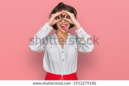 Young hispanic woman wearing business style and glasses doing ok gesture like binoculars sticking tongue out, eyes looking through fingers. crazy expression. 