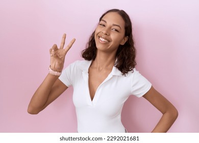 Young hispanic woman wearing casual white t shirt smiling looking to the camera showing fingers doing victory sign. number two. 