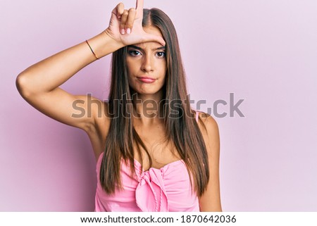 Young hispanic woman wearing casual style with sleeveless shirt making fun of people with fingers on forehead doing loser gesture mocking and insulting. 
