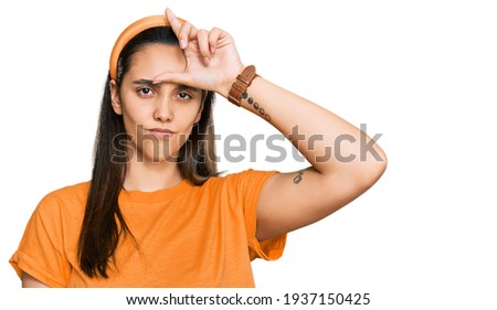 Young hispanic woman wearing casual clothes making fun of people with fingers on forehead doing loser gesture mocking and insulting. 