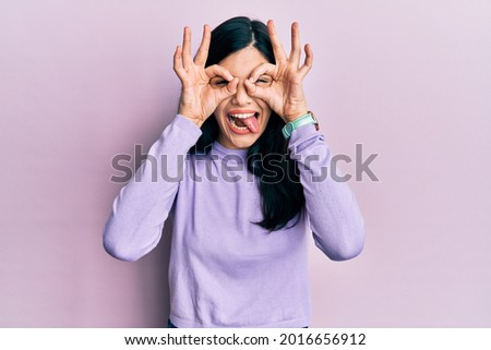 Young hispanic woman wearing casual clothes doing ok gesture like binoculars sticking tongue out, eyes looking through fingers. crazy expression. 