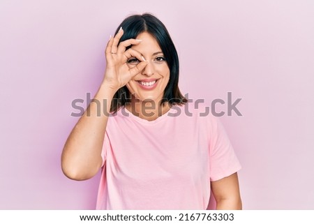 Young hispanic woman wearing casual pink t shirt doing ok gesture with hand smiling, eye looking through fingers with happy face. 