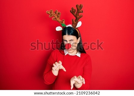 Young hispanic woman wearing deer christmas hat and red nose disgusted expression, displeased and fearful doing disgust face because aversion reaction. 
