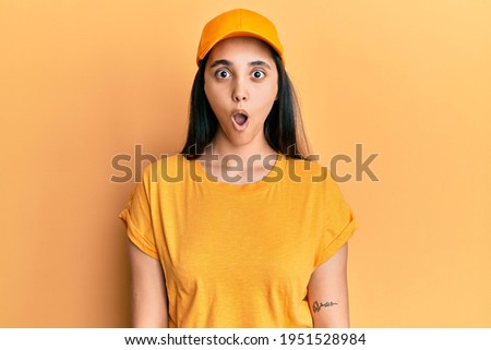 Young hispanic woman wearing delivery uniform and cap afraid and shocked with surprise expression, fear and excited face. 