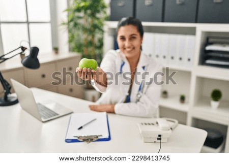 Young hispanic woman wearing dietitian uniform holding apple at clinic