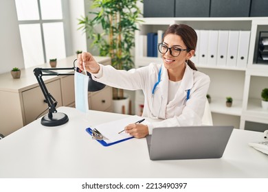 Young Hispanic Woman Wearing Doctor Uniform Holding Medical Mask At Clinic