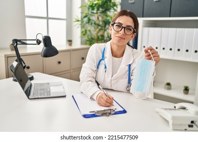 Young Hispanic Woman Wearing Doctor Uniform Holding Medical Mask At Clinic