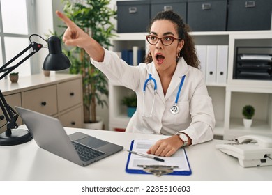 Young hispanic woman wearing doctor uniform and stethoscope pointing with finger surprised ahead, open mouth amazed expression, something on the front  - Shutterstock ID 2285510603