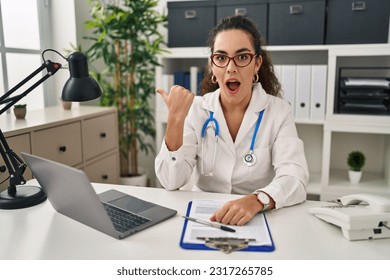 Young hispanic woman wearing doctor uniform and stethoscope surprised pointing with hand finger to the side, open mouth amazed expression.  - Shutterstock ID 2317265785