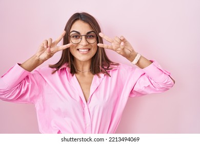 Young Hispanic Woman Wearing Glasses Standing Over Pink Background Doing Peace Symbol With Fingers Over Face, Smiling Cheerful Showing Victory 