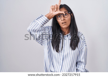 Young hispanic woman wearing glasses making fun of people with fingers on forehead doing loser gesture mocking and insulting. 