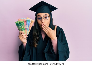 Young hispanic woman wearing graduation uniform holding south africa rand banknotes covering mouth with hand, shocked and afraid for mistake. surprised expression 