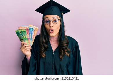 Young hispanic woman wearing graduation uniform holding south africa rand banknotes scared and amazed with open mouth for surprise, disbelief face 
