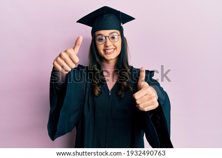 Young hispanic woman wearing graduation cap and ceremony robe approving doing positive gesture with hand, thumbs up smiling and happy for success. winner gesture. 