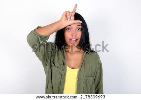 Young hispanic woman wearing green jacket over white background gestures with finger on forehead makes loser gesture makes fun of people shows tongue