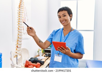 Young hispanic woman wearing physiotherapist uniform pointing to vertebral column using touchpad at physiotherapy clinic