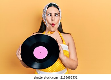 Young Hispanic Woman Wearing Pin Up Style Holding Vinyl Disc Making Fish Face With Mouth And Squinting Eyes, Crazy And Comical. 