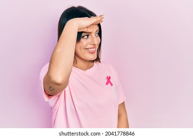 Young Hispanic Woman Wearing Pink Cancer Ribbon On T Shirt Very Happy And Smiling Looking Far Away With Hand Over Head. Searching Concept. 