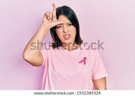 Young hispanic woman wearing pink cancer ribbon on t shirt making fun of people with fingers on forehead doing loser gesture mocking and insulting. 