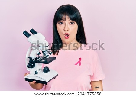 Young hispanic woman wearing pink cancer ribbon on shirt holding microscope scared and amazed with open mouth for surprise, disbelief face 