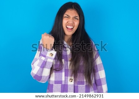young hispanic woman wearing plaid shirt over blue background angry and mad raising fist frustrated and furious while shouting with anger. Rage and aggressive concept.