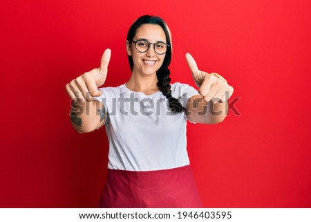 Young hispanic woman wearing professional waitress apron approving doing positive gesture with hand, thumbs up smiling and happy for success. winner gesture. 