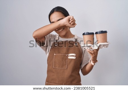 Young hispanic woman wearing professional waitress apron holding coffee smiling cheerful playing peek a boo with hands showing face. surprised and exited 