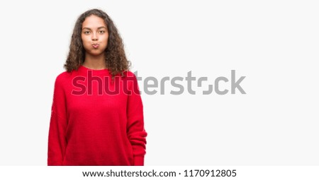 Young hispanic woman wearing red sweater puffing cheeks with funny face. Mouth inflated with air, crazy expression.