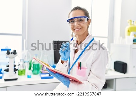 Young hispanic woman wearing scientist uniform writing on notebook at laboratory
