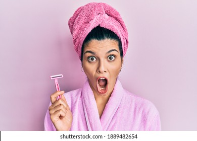 Young hispanic woman wearing shower bathrobe holding razor scared and amazed with open mouth for surprise, disbelief face 