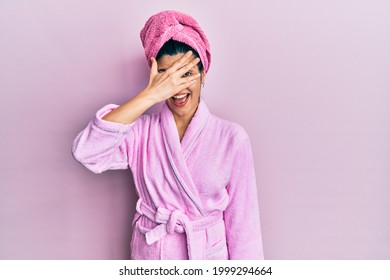 Young Hispanic Woman Wearing Shower Towel Cap And Bathrobe Peeking In Shock Covering Face And Eyes With Hand, Looking Through Fingers With Embarrassed Expression. 