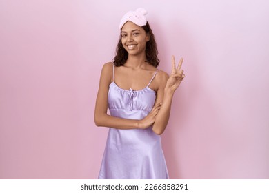 Young hispanic woman wearing sleep mask and nightgown smiling with happy face winking at the camera doing victory sign with fingers. number two. 