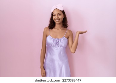 Young hispanic woman wearing sleep mask and nightgown smiling cheerful presenting and pointing with palm of hand looking at the camera. 
