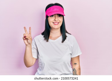 Young hispanic woman wearing sportswear and sun visor cap smiling with happy face winking at the camera doing victory sign. number two. 