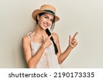 Young hispanic woman wearing summer hat smiling and looking at the camera pointing with two hands and fingers to the side. 