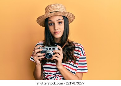 Young Hispanic Woman Wearing Summer Hat Holding Vintage Camera Relaxed With Serious Expression On Face. Simple And Natural Looking At The Camera. 