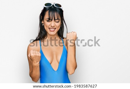Young hispanic woman wearing swimsuit and sunglasses celebrating surprised and amazed for success with arms raised and eyes closed. winner concept. 