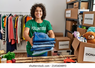 Young hispanic woman wearing volunteer uniform holding folded jeans at charity center