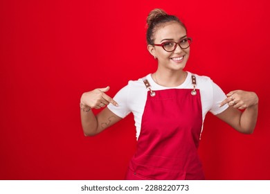 Young hispanic woman wearing waitress apron over red background looking confident with smile on face, pointing oneself with fingers proud and happy.  - Powered by Shutterstock